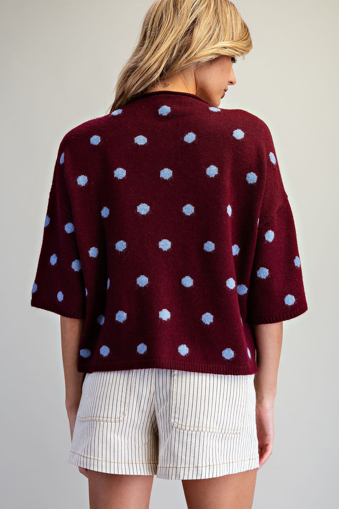 A woman shown from the back wearing a burgundy knit cardigan with light blue polka dots and cream pinstriped shorts against a plain light gray background.