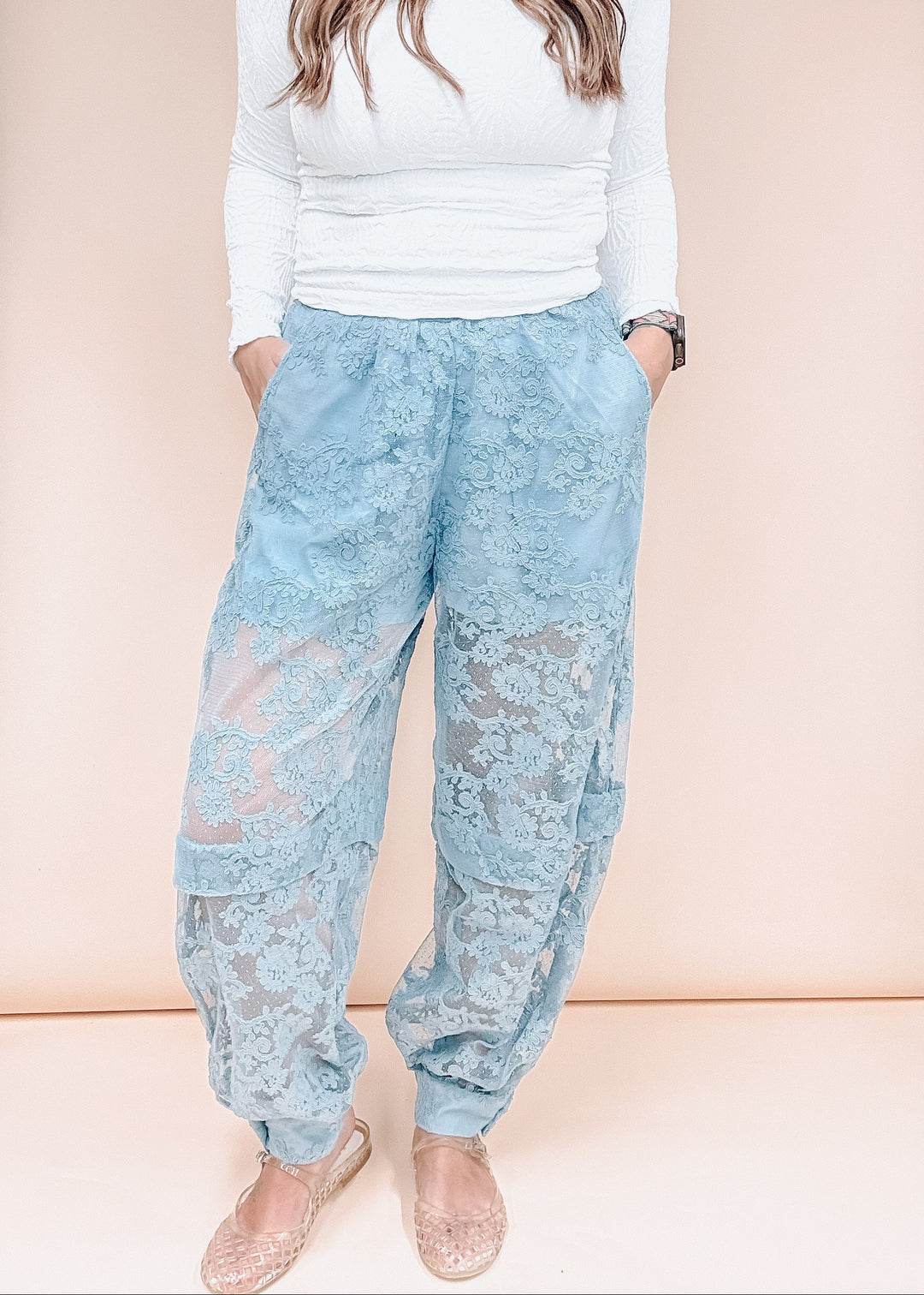 A model wearing light blue lace harem pants with a white long-sleeve top and beige flat shoes, standing against a solid pale peach background.