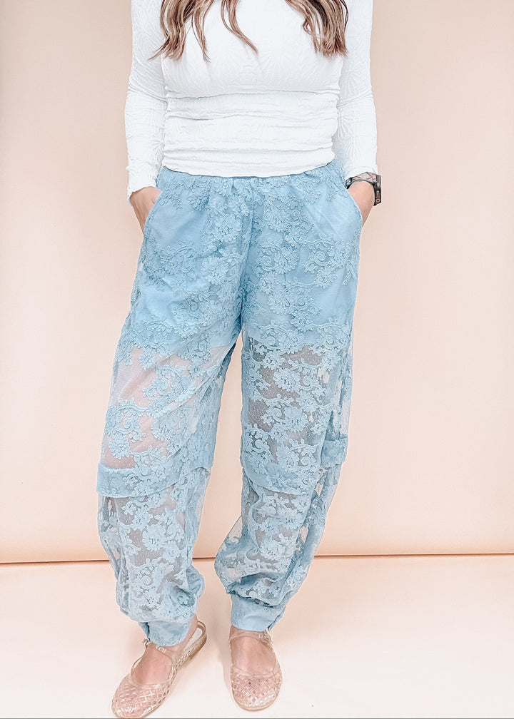A model wearing light blue lace harem pants with a white long-sleeve top and beige flat shoes, standing against a solid pale peach background.