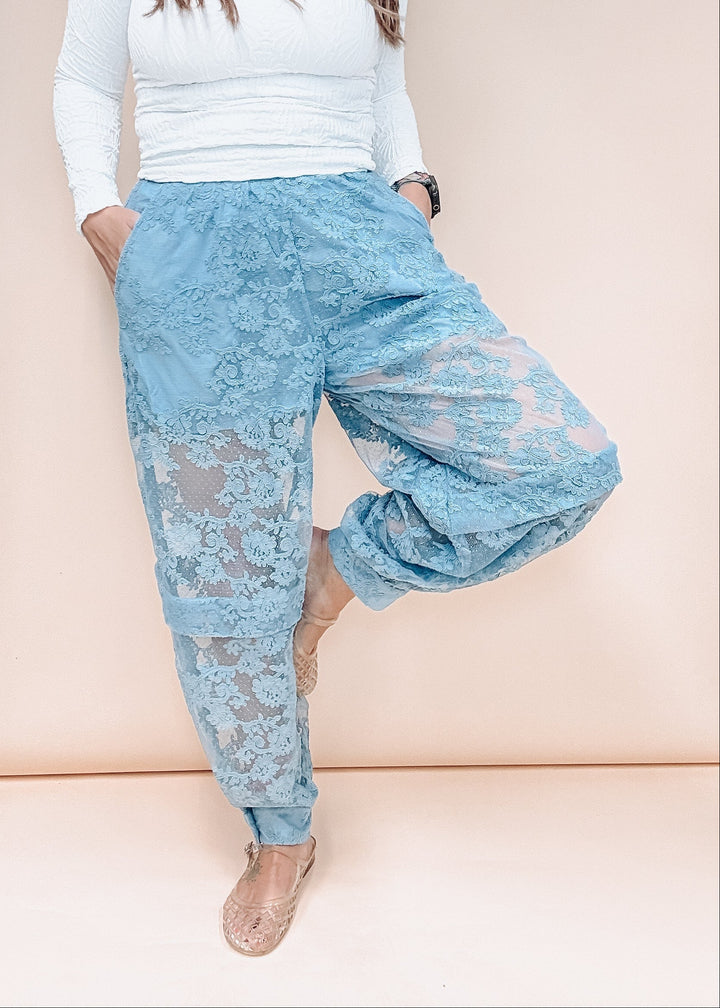 A woman models light blue lace harem-style pants with a white ribbed long-sleeve top and beige flat shoes, standing against a solid pale peach background.