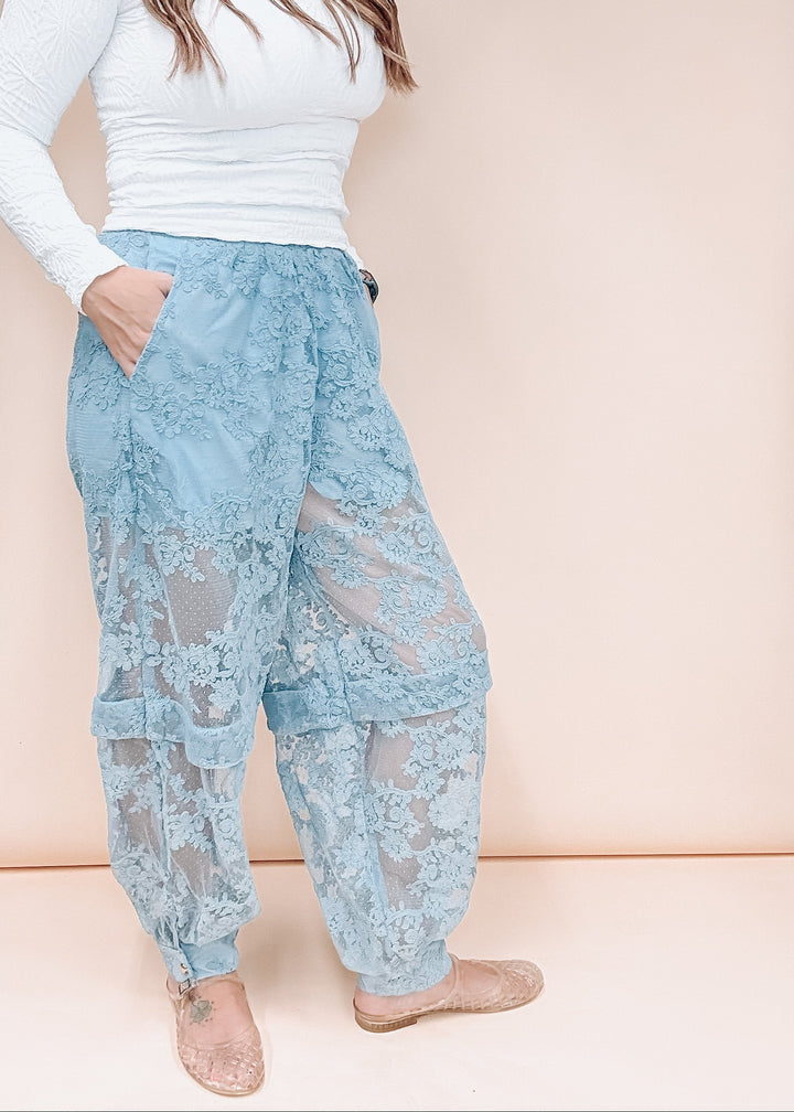 A model wearing light blue lace harem-style pants with a white long-sleeve top and nude flat shoes, standing against a solid pale peach background.