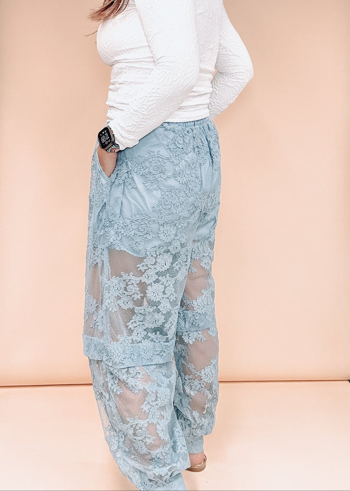 A person wearing light blue lace wide-leg pants with sheer mesh panels and a white textured long-sleeve top, standing against a soft peach studio backdrop.