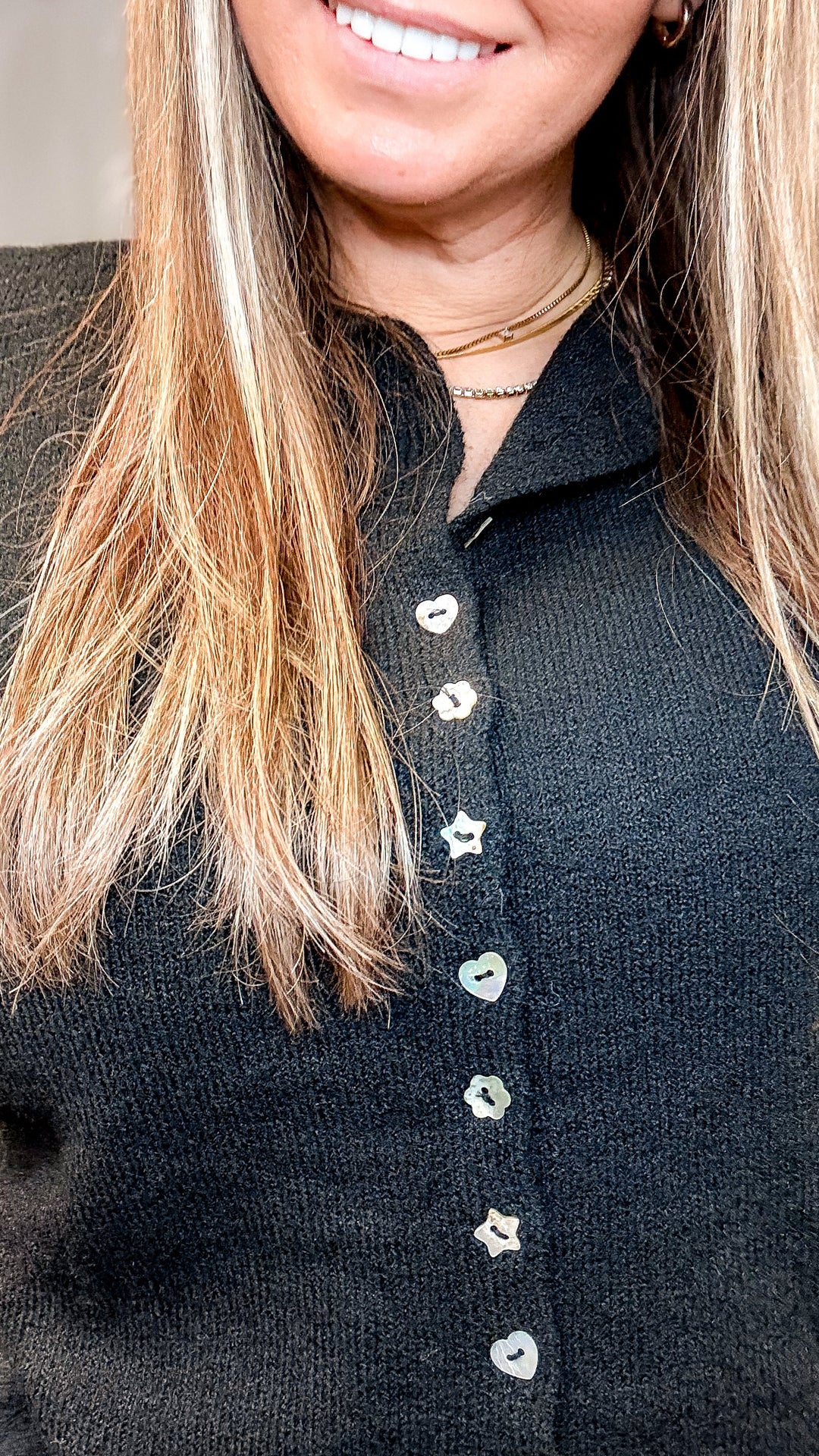 Close-up of a woman wearing a black knit button-up top with white star-shaped buttons and layered gold necklaces.