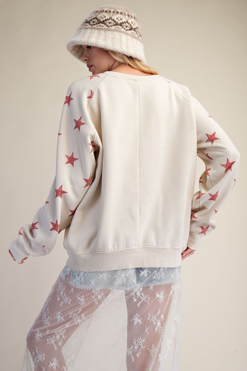 Person wearing a white sweater with red star patterns and a patterned hat on a beige background