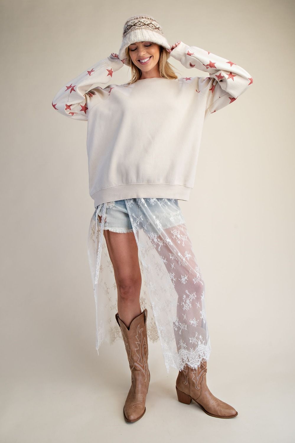 Woman wearing a white sweater with floral sleeves, denim shorts, and brown cowboy boots on a plain background