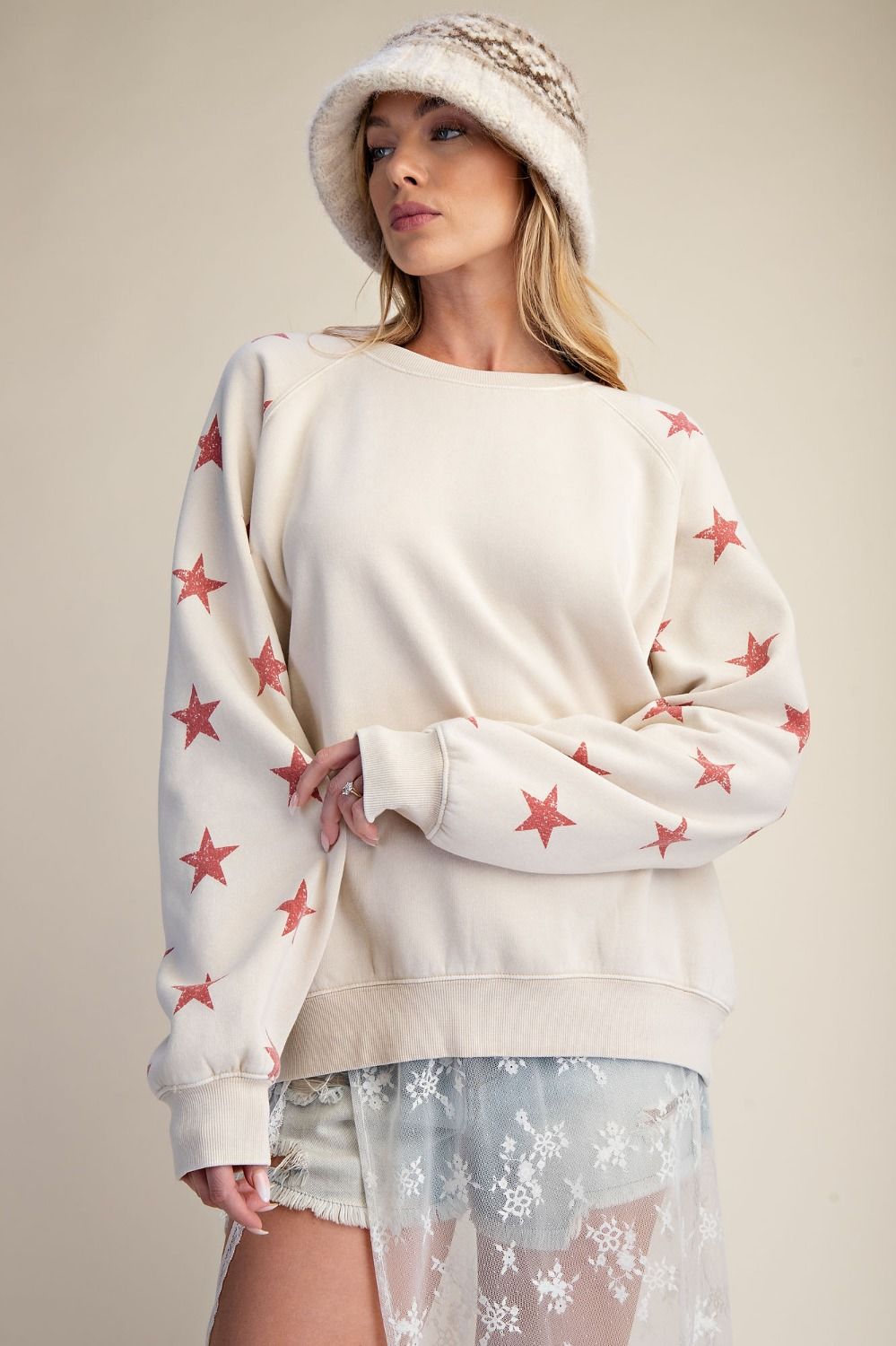 Woman wearing a cream sweater with red star patterns and a white hat against a neutral background