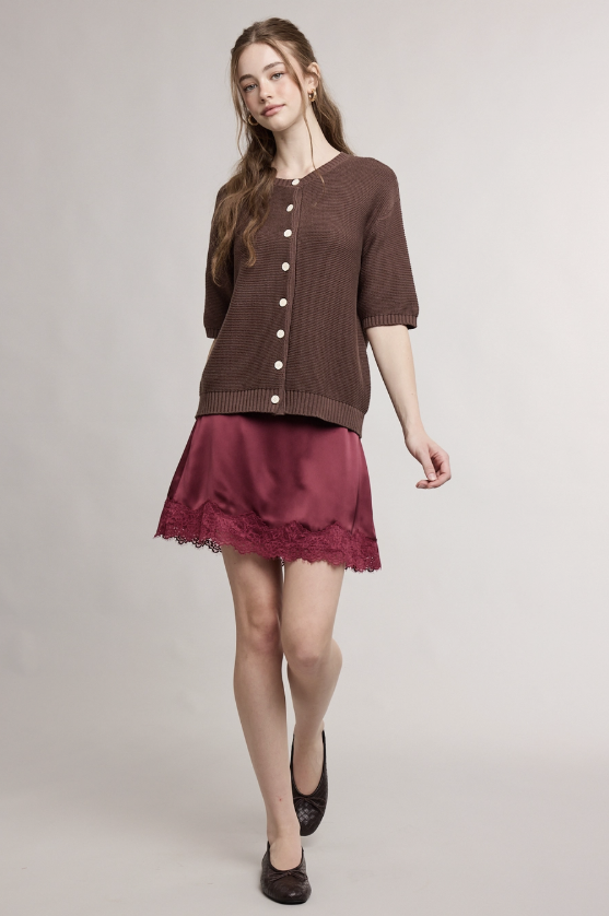 A woman modeling a brown short-sleeve button-front knit cardigan over a burgundy satin mini skirt with a lace hem, paired with black flats, against a light gray studio background.