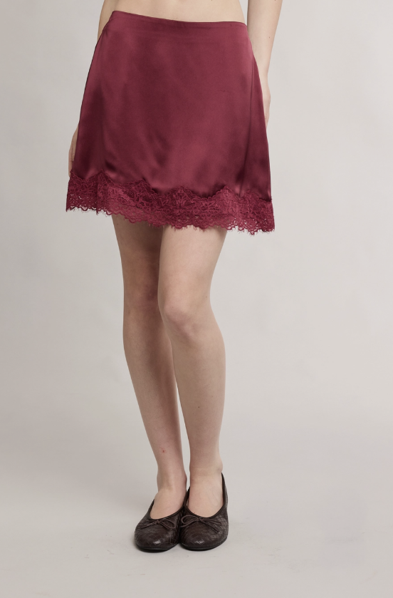 A person wearing a short satin-like burgundy skirt with a lace hem, paired with dark flat shoes, photographed against a plain light gray studio background.