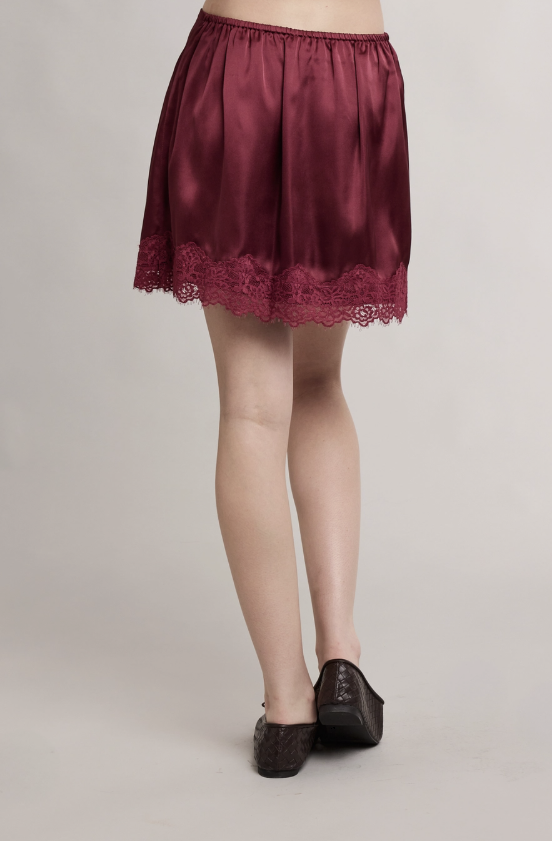 A person wearing a short satin skirt in deep burgundy with a lace hem, shown from the waist down against a plain light gray studio background.