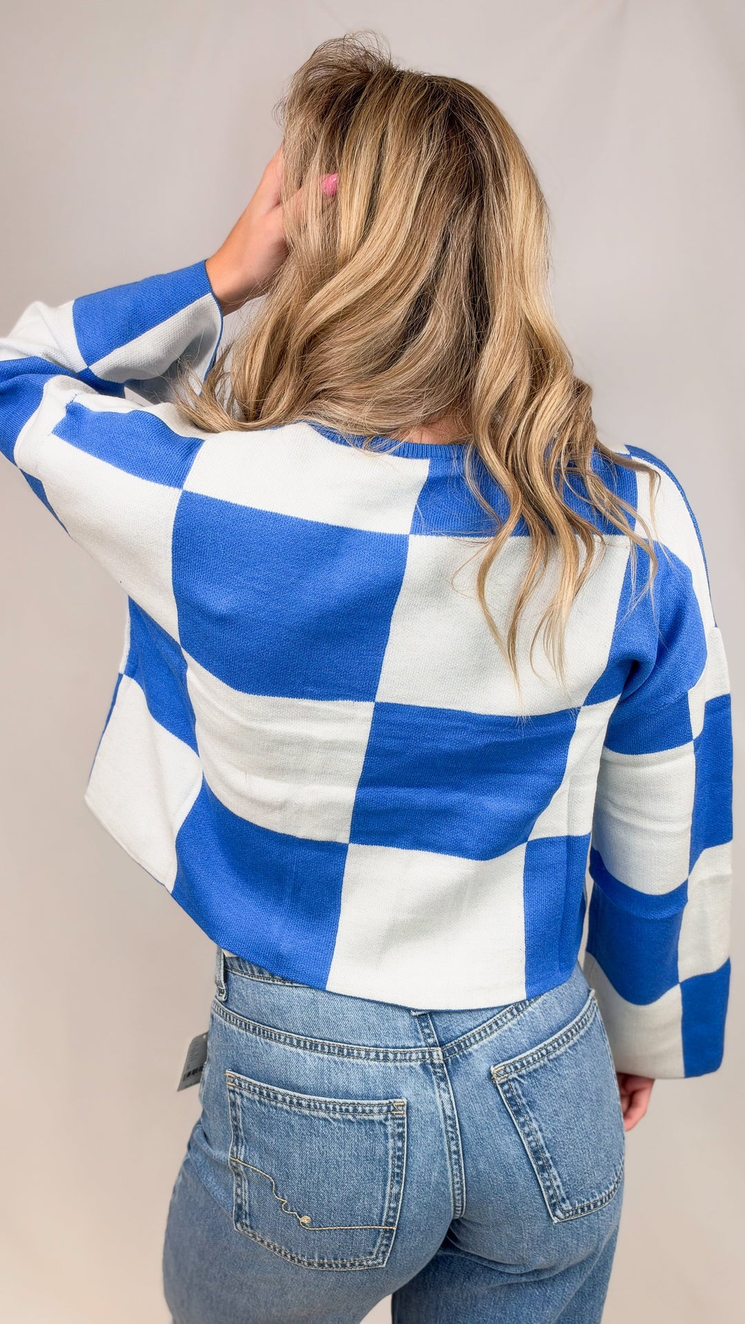 Woman shown from back wearing blue and white checker crop sweater with jeans on plain background