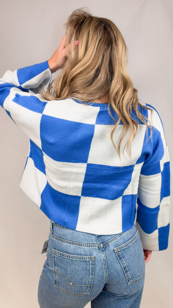 Woman shown from back wearing blue and white checker crop sweater with jeans on plain background