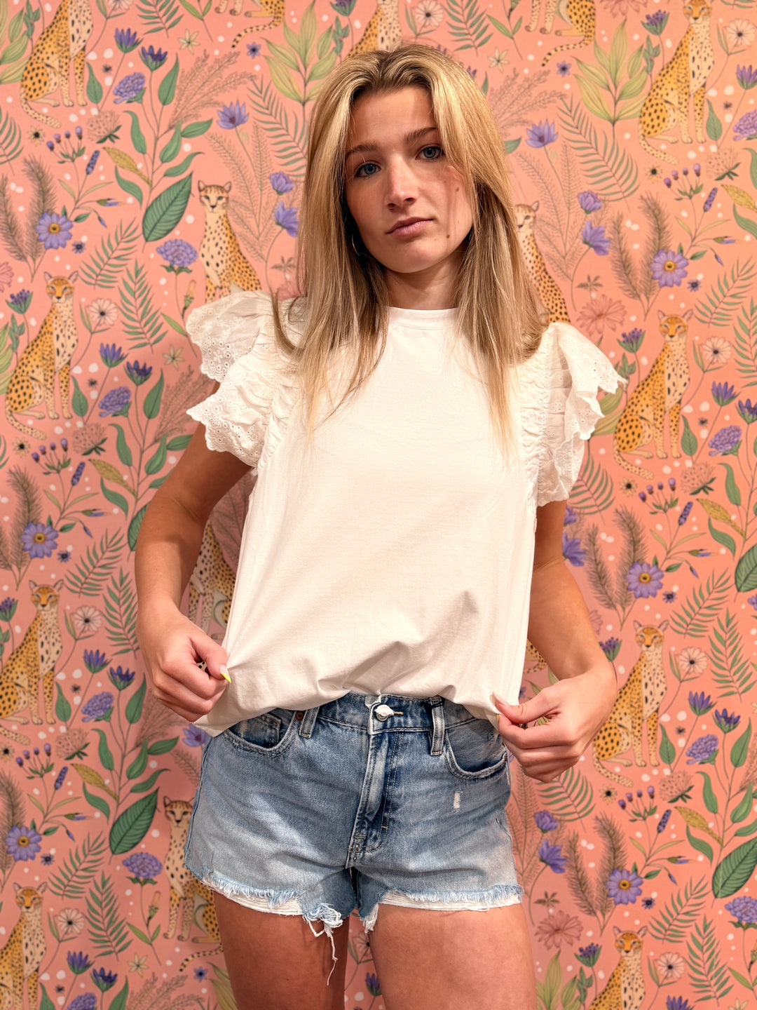 Woman wearing a white ruffled blouse and denim shorts against a pink floral and animal patterned background