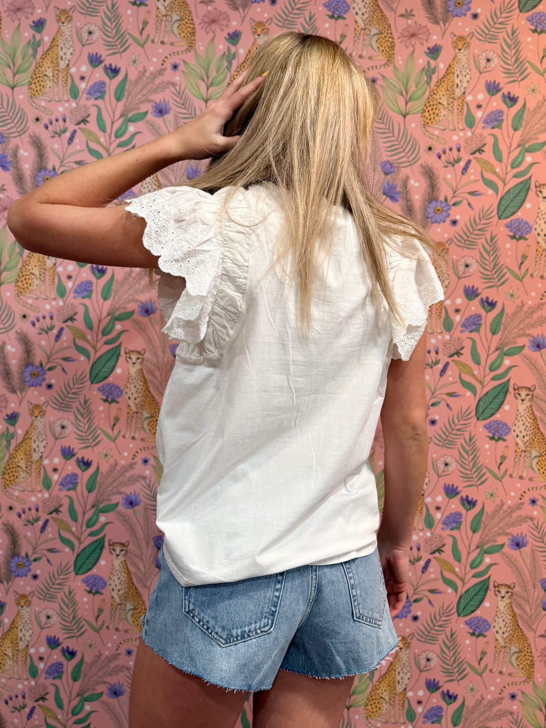 Person wearing a white blouse with ruffled sleeves and denim shorts against a pink floral and cheetah patterned wall.