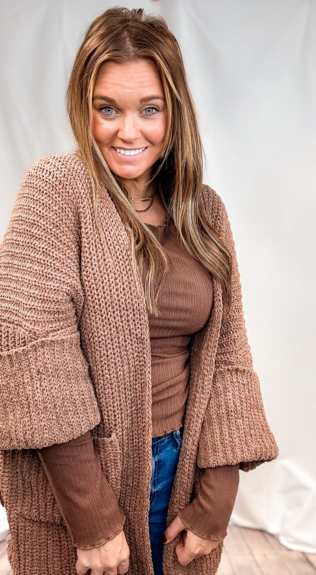 A smiling woman with long light brown hair wearing a chunky, open-front brown knit cardigan over a brown ribbed top and blue jeans, standing against a white fabric backdrop.