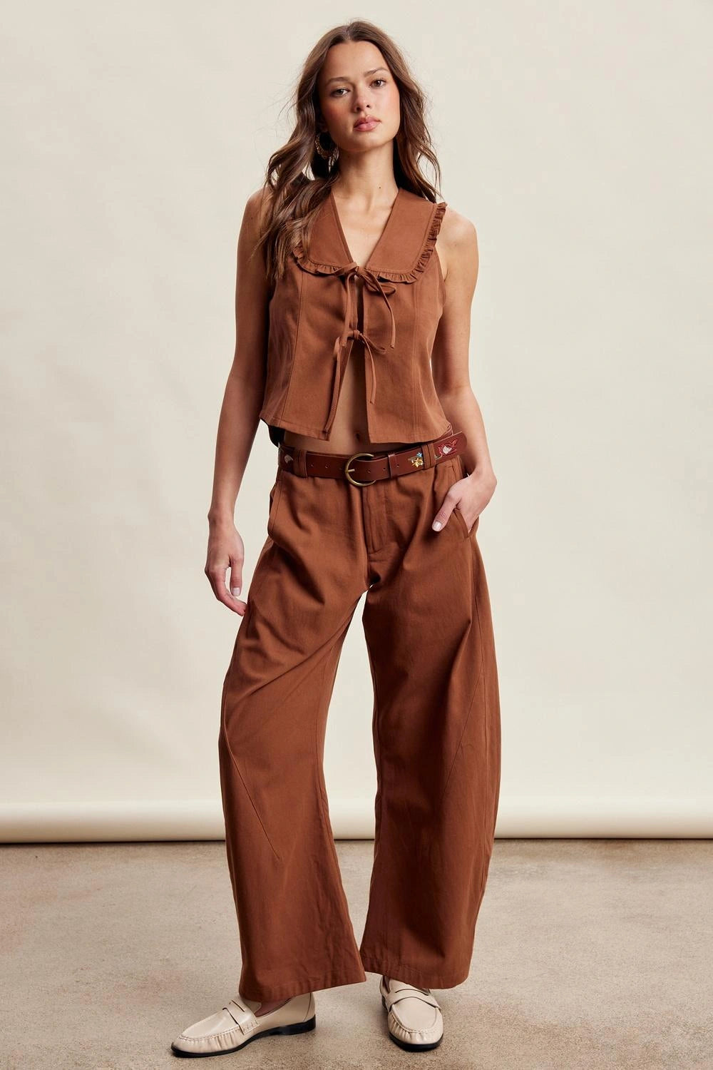 A woman modeling a rust-brown two-piece set consisting of a sleeveless tie-front top and matching wide-leg pants, styled with a matching belt and white loafers against a neutral studio backdrop.