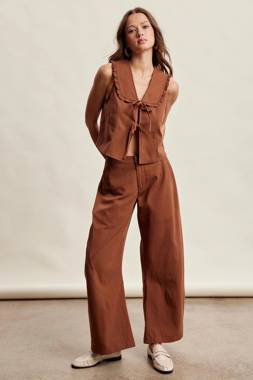 A woman modeling a rust-brown two-piece outfit consisting of a sleeveless ruffled tie-front top and matching wide-leg pants, paired with white slip-on shoes against a plain studio backdrop.