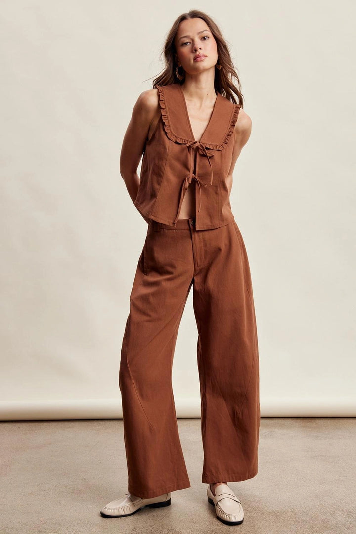 A woman modeling a rust-brown two-piece outfit consisting of a sleeveless ruffled tie-front top and matching wide-leg pants, paired with white slip-on shoes against a plain studio backdrop.