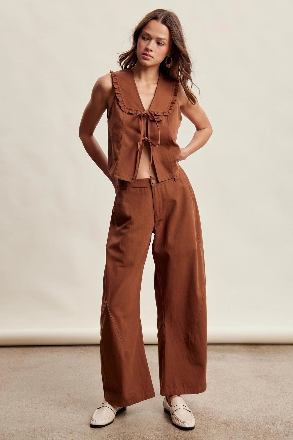 A woman modeling a rust-brown two-piece linen set consisting of a sleeveless tie-front top and matching wide-leg pants, paired with white loafers, against a neutral studio backdrop.
