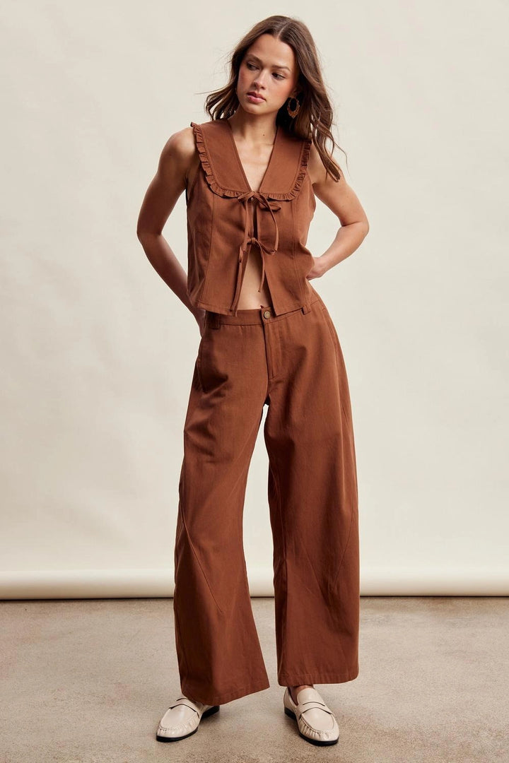 A woman modeling a rust-brown two-piece linen set consisting of a sleeveless tie-front top and matching wide-leg pants, paired with white loafers, against a neutral studio backdrop.