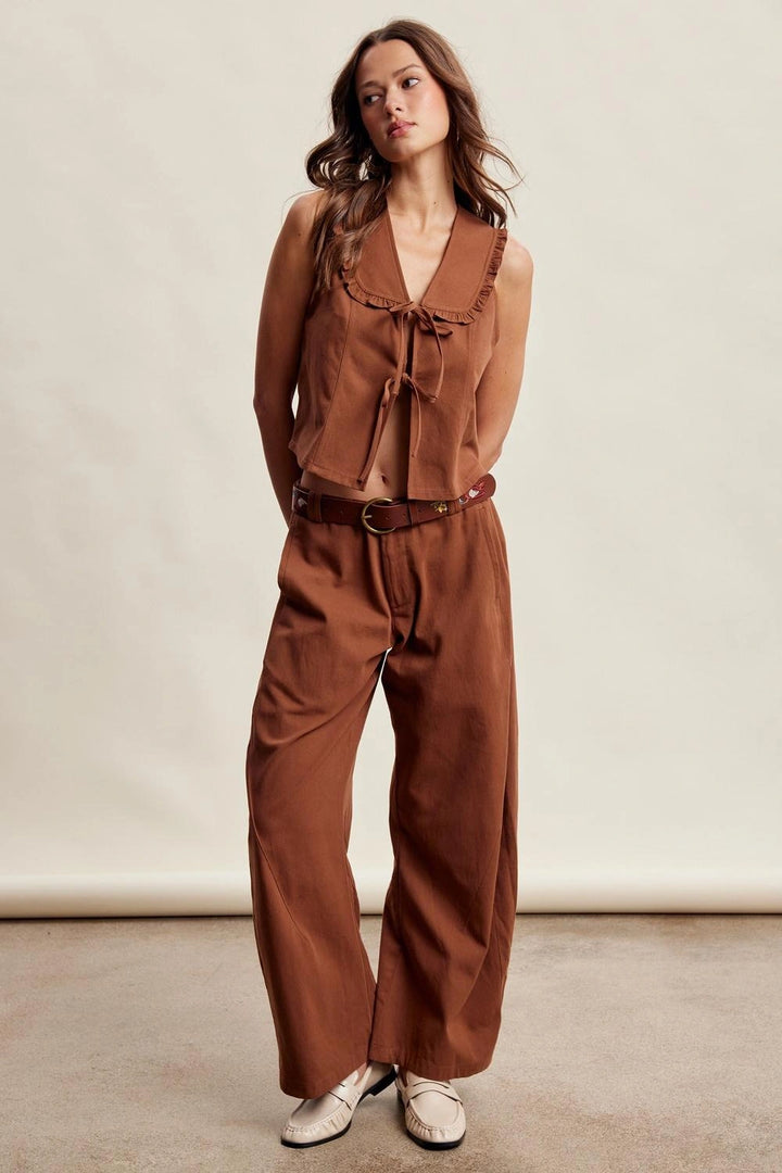 A woman modeling a rust-brown two-piece outfit consisting of a sleeveless ruffled tie-front top and matching wide-leg pants, styled with a brown belt and white shoes against a plain beige studio backdrop.