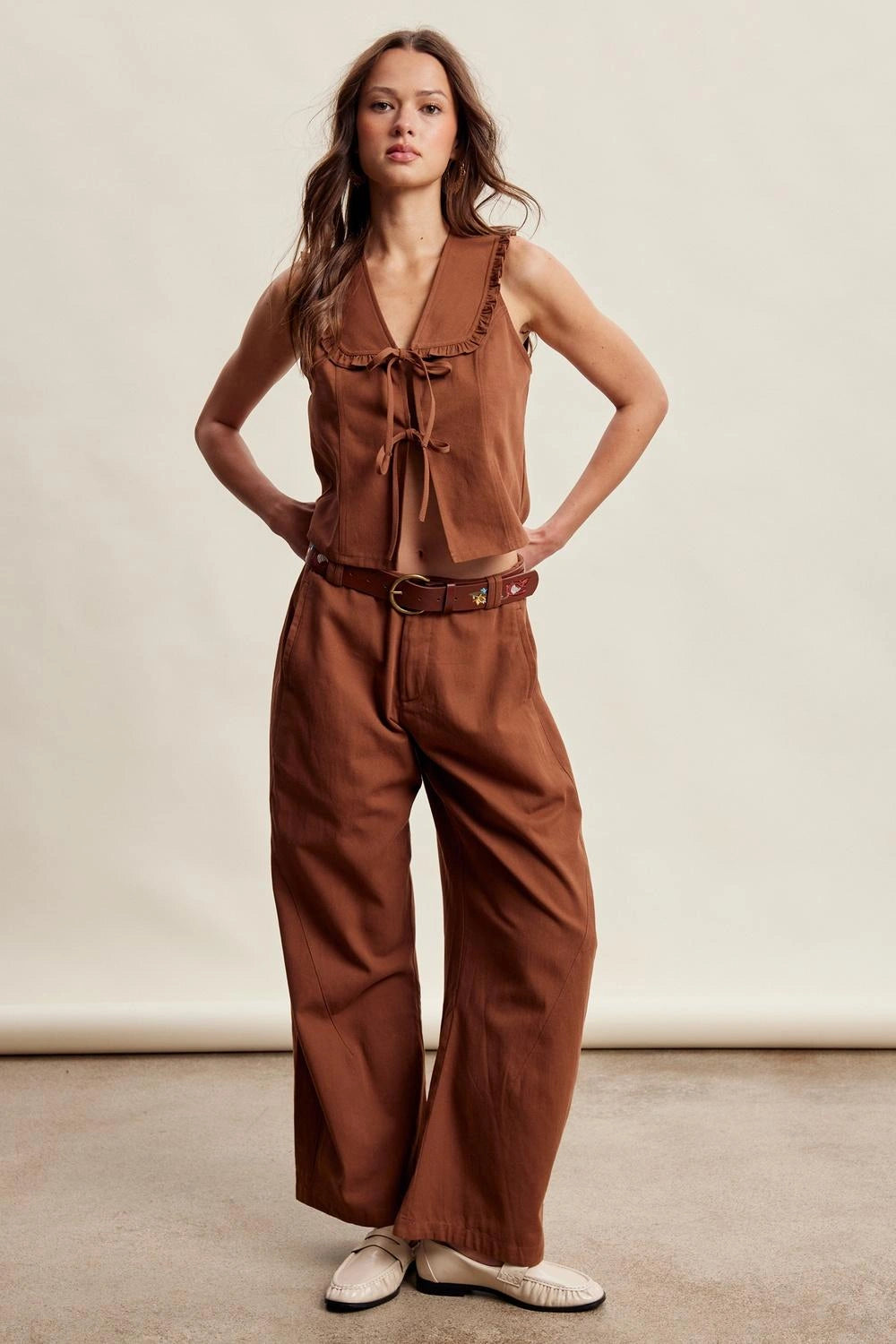 A woman modeling a rust-brown sleeveless jumpsuit with a tie-neck detail, wide legs, and a matching brown belt, paired with white slip-on shoes against a neutral studio backdrop.