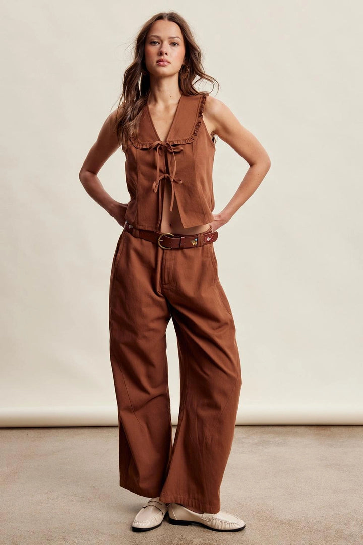 A woman modeling a rust-brown sleeveless jumpsuit with a tie-neck detail, wide legs, and a matching brown belt, paired with white slip-on shoes against a neutral studio backdrop.