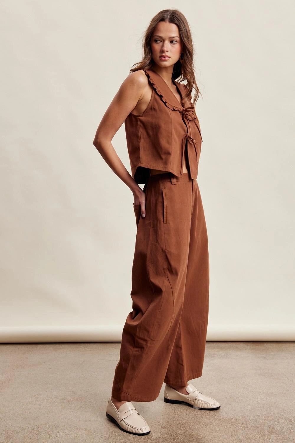 A woman modeling a rust-brown two-piece linen set consisting of a sleeveless ruffle-trimmed crop top and matching wide-leg trousers, paired with cream-colored loafers against a neutral studio backdrop.