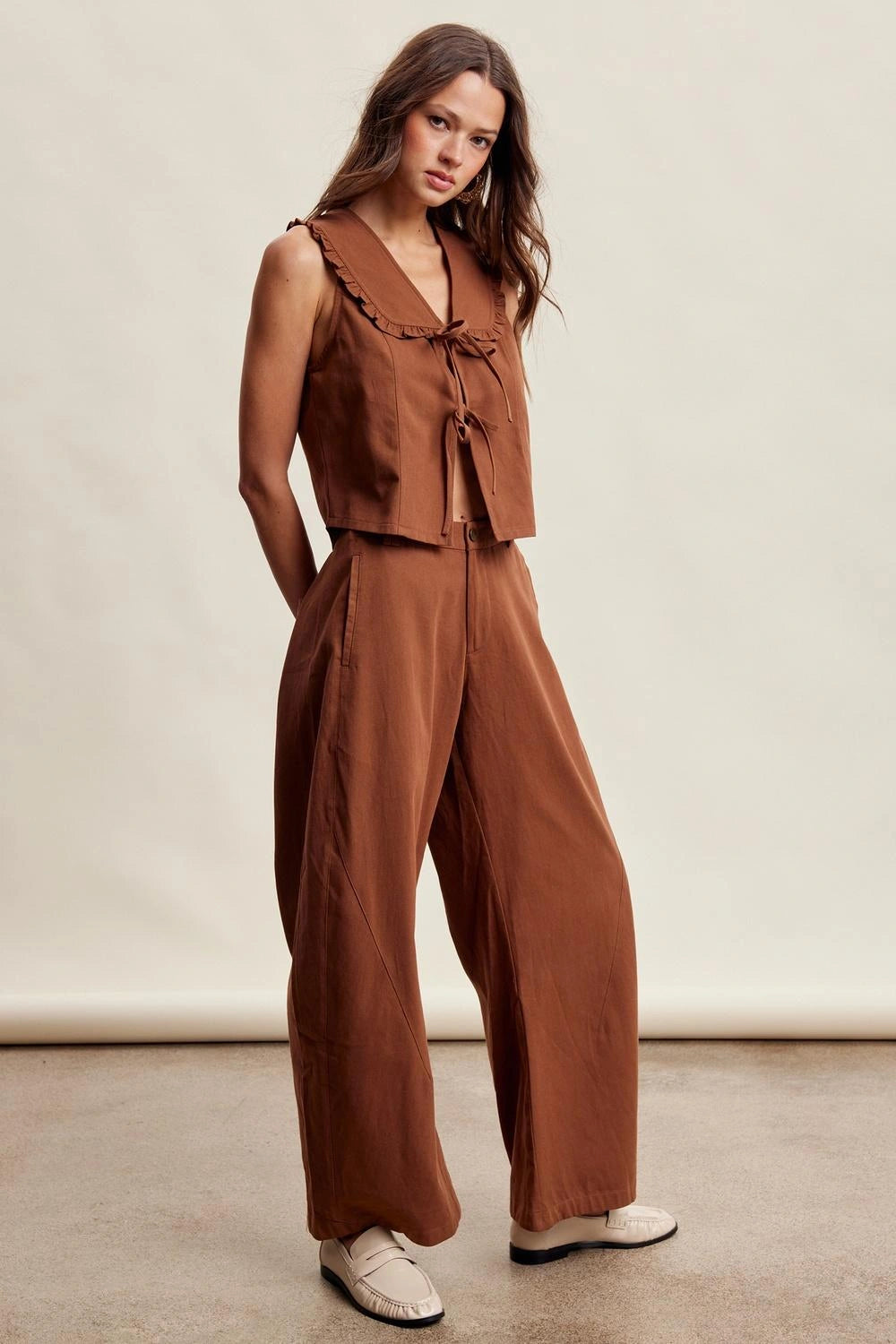 A woman modeling a rust-brown two-piece linen set consisting of a sleeveless cropped top with ruffle trim and tie details, paired with matching wide-leg trousers, against a neutral studio backdrop.
