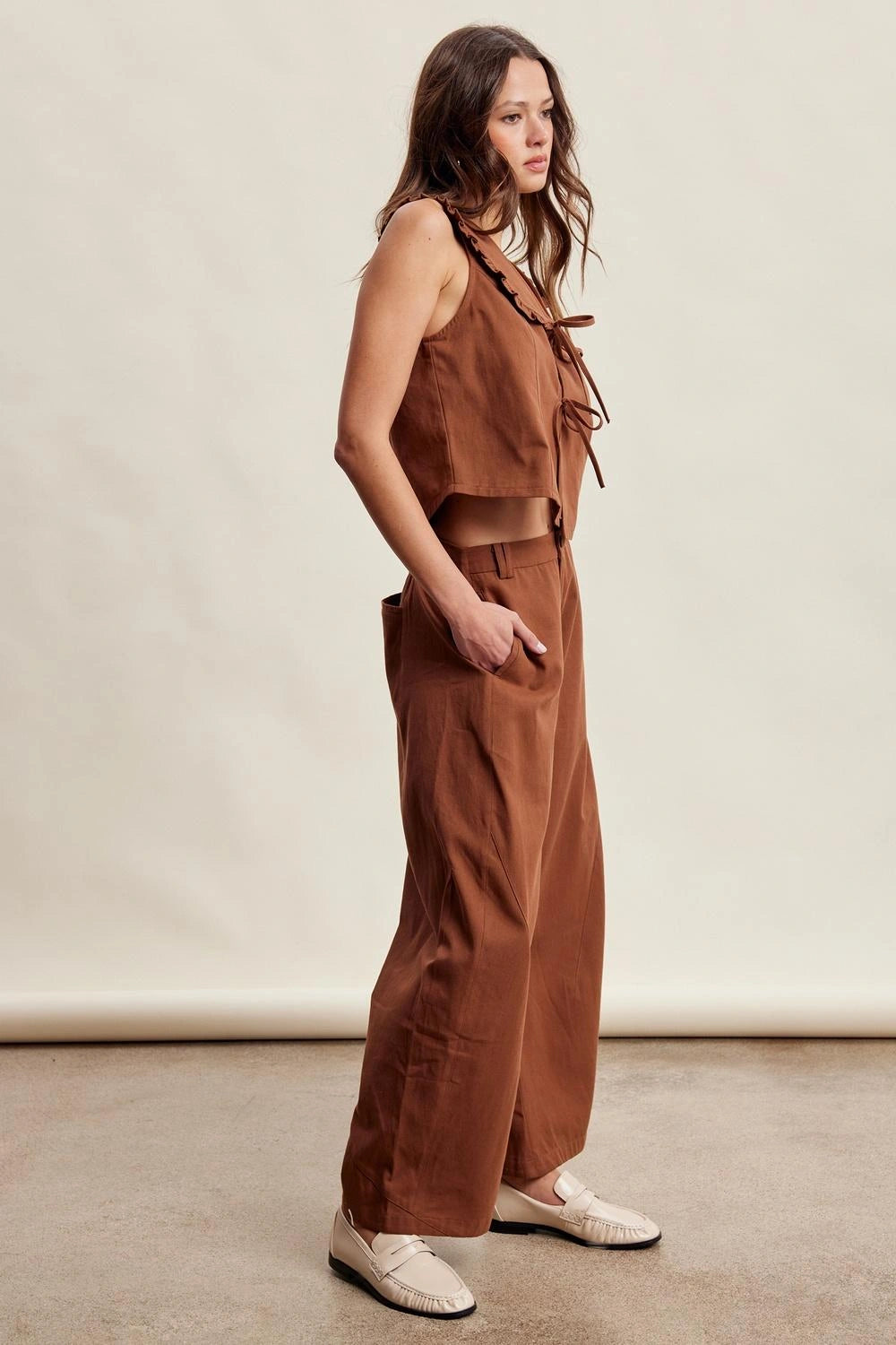 A woman models a rust-brown two-piece linen set consisting of a sleeveless crop top with tie details and matching wide-leg pants, paired with cream-colored loafers, against a neutral studio backdrop.