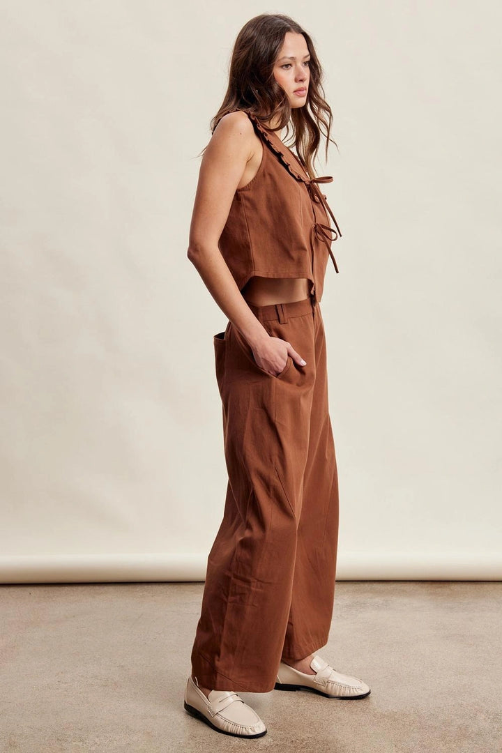 A woman models a rust-brown two-piece linen set consisting of a sleeveless crop top with tie details and matching wide-leg pants, paired with cream-colored loafers, against a neutral studio backdrop.