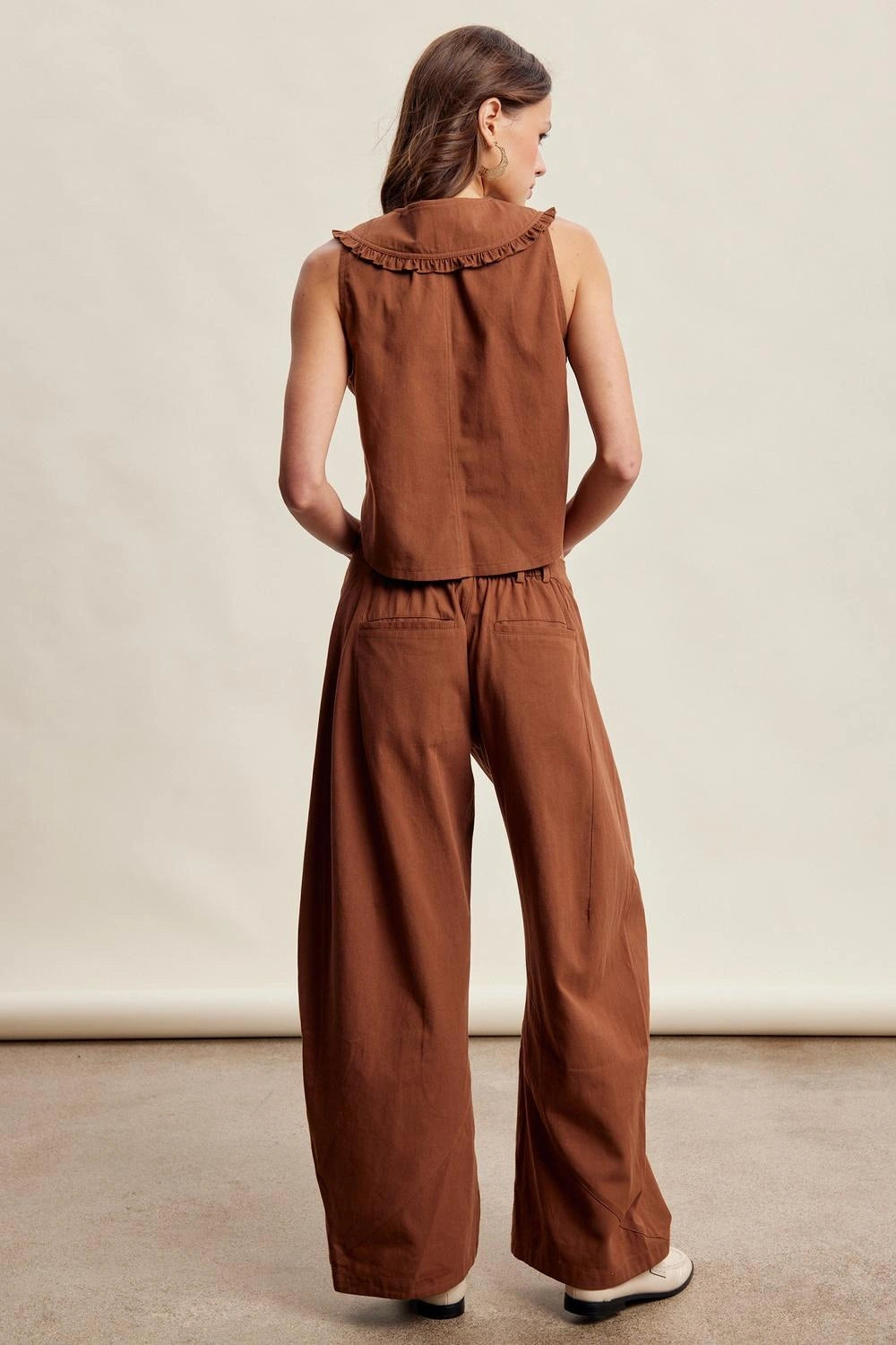A woman shown from the back wearing a rust-brown sleeveless top with a ruffled collar and matching wide-leg pants, photographed in a studio setting.