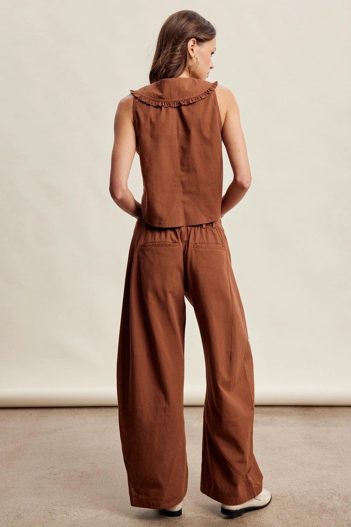 A woman shown from the back wearing a rust-brown sleeveless top with a ruffled collar and matching wide-leg pants, photographed in a studio setting.