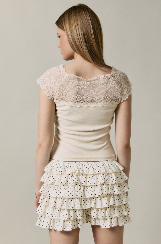 A woman shown from the back wearing a cream-colored top with a lace yoke and a matching cream ruffled mini skirt with small black polka dots, set against a plain light gray studio background.