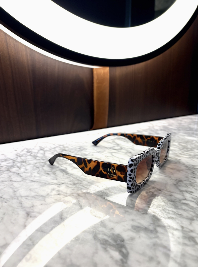 A pair of square sunglasses with a leopard print frame and patterned temples, resting on a white marble surface in front of a dark wood background with a circular light fixture.
