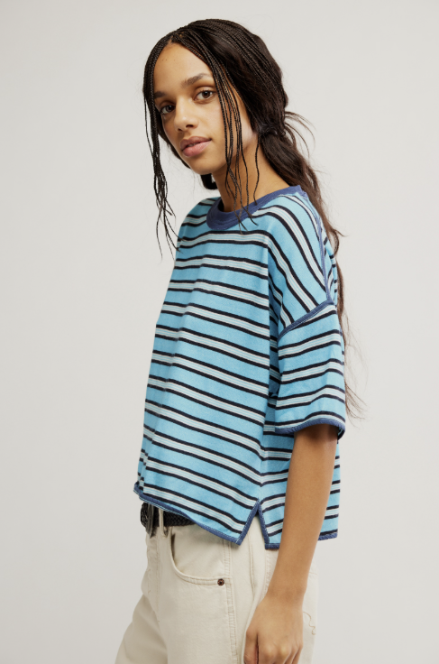 A model wearing a light blue and navy striped short-sleeve crop top with a relaxed fit, paired with off-white jeans, against a plain light gray studio background.