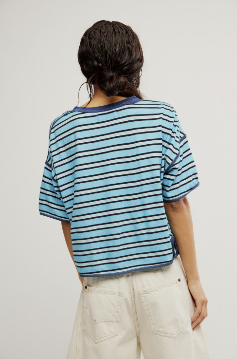 A person shown from the back wearing a light blue and navy striped short-sleeve t-shirt and off-white pants, against a plain light gray studio background.