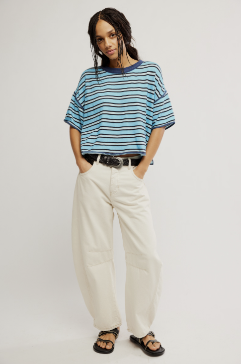 A woman modeling a blue and navy striped short-sleeve t-shirt, paired with cream wide-leg trousers, a black belt, and black sandals, against a plain light gray studio background.