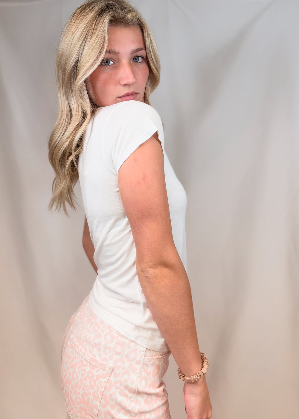Woman shown from the side wearing white baby tee with pink leopard pants on plain backgorund