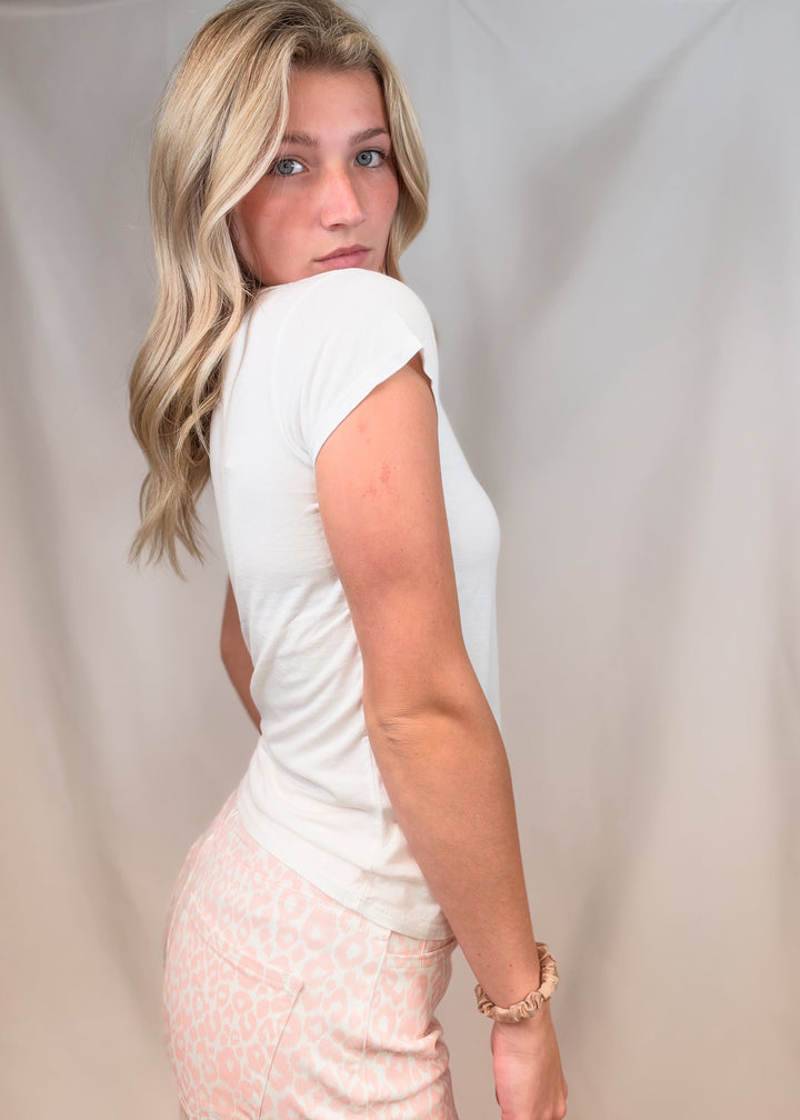 Woman shown from the side wearing white baby tee with pink leopard pants on plain backgorund
