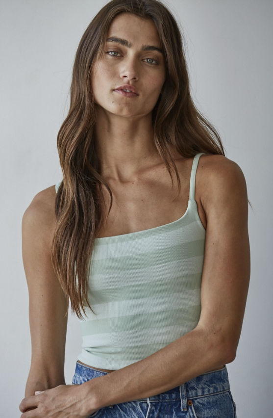 A woman wearing a pale sage green and white horizontally striped camisole tank top with blue jeans, photographed against a neutral light gray background.