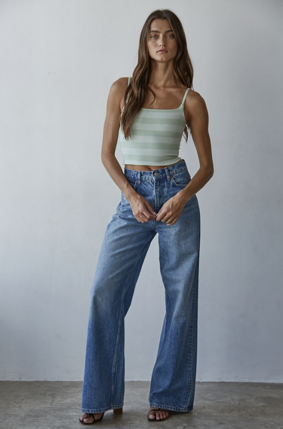 A woman modeling a light green and white striped camisole top with high-waisted, wide-leg blue jeans against a plain light gray wall.