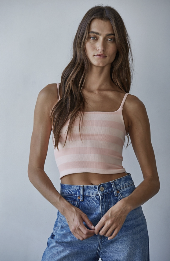 A woman modeling a light pink and white horizontally striped spaghetti-strap crop top paired with high-waisted blue jeans, set against a plain light gray studio background.