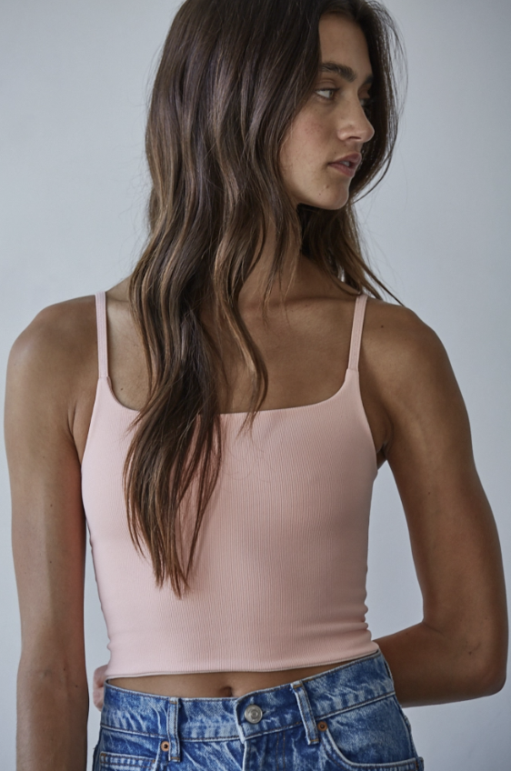 A woman wearing a light pink spaghetti-strap crop top and blue denim jeans, shown from the waist up against a plain light gray studio background.
