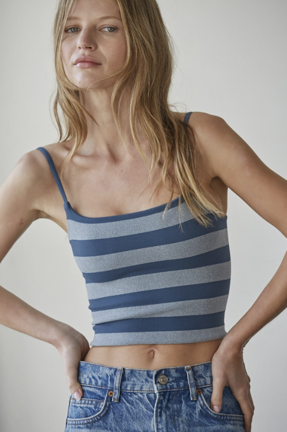 A woman wearing a blue and gray striped crop top with thin straps, paired with blue jeans, photographed against a plain light gray background.