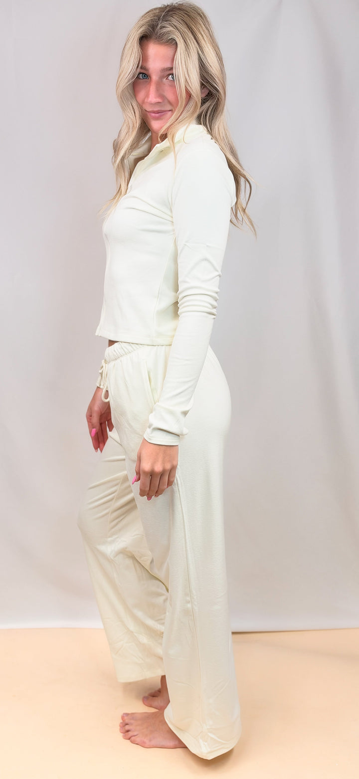 A woman modeling a cream-colored two-piece loungewear set consisting of a long-sleeve cropped top and matching wide-leg pants, standing against a light beige studio backdrop.