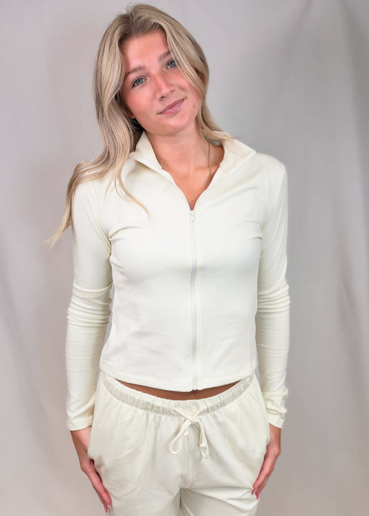 Woman wearing cream color matching set with zip up top and pants on plain background