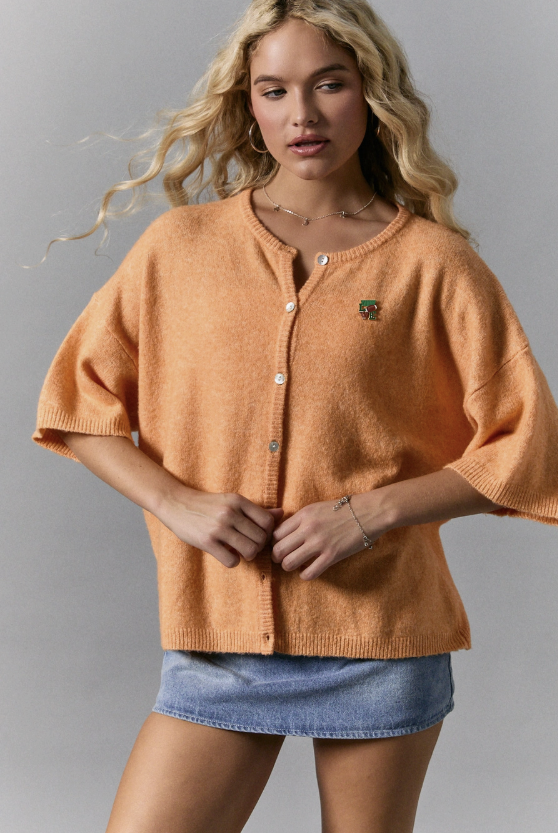 Woman wearing an orange sweater and denim shorts against a gray background