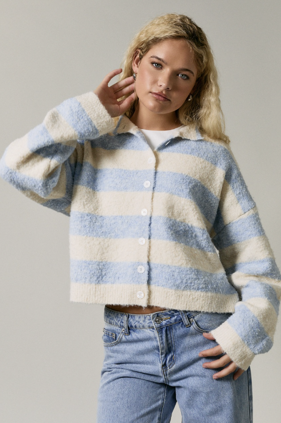 A woman wearing a fuzzy, cropped button-up cardigan with horizontal light blue and cream stripes, paired with light-wash blue jeans, against a plain light gray studio background.