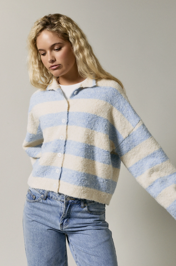 A woman wearing a fuzzy, cropped button-up cardigan with horizontal light blue and cream stripes, paired with light-wash blue jeans, photographed against a neutral light gray studio background.