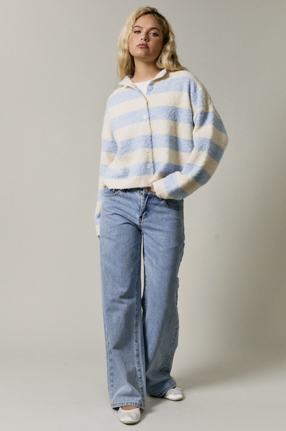 A woman modeling a fuzzy, light blue and cream striped cropped cardigan sweater with wide-leg light blue jeans and white shoes, standing against a neutral light gray studio background.